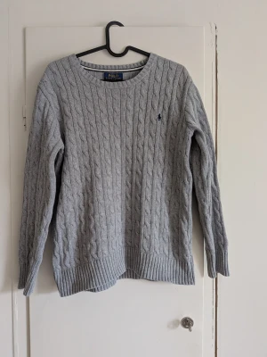 Grå kabelstickad tröja Polo Ralph Lauren - Kabelstickad grå tröja från Polo Ralph Lauren med rund halsringning och diskret broderad logga på bröstet. Tröjan har långa ärmar och ribbade muddar vid ärmslut och nederkant. Perfekt för lager-på-lager och klassisk stil.