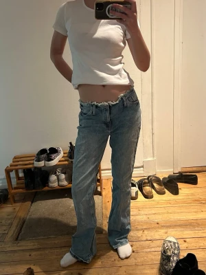 Ljusblå jeans med rå kant - Säljer ett par ljusblå jeans med rå, fransad midja och klassisk femficksdesign. Jeansen har en avslappnad passform och lätt utsvängda ben. Perfekta för en trendig och avslappnad look.
