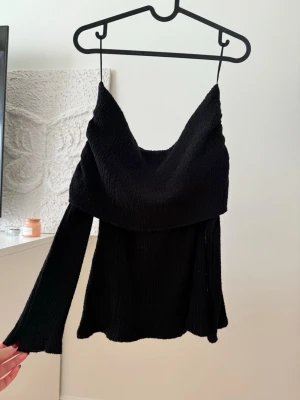 Svart offshoulder topp med vida ärmar - Trendig svart offshoulder topp med vida långa ärmar och snygg draperad krage. Toppens material är strukturerat och ger en cool känsla. Perfekt för dig som vill sticka ut med en stilren och modern look.