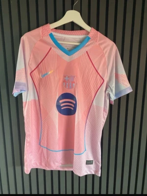 Barça rosa och blå matchtröja - Säljer en FC Barcelona matchtröja i rosa och ljusblått med klubbmärke och Spotify-logga på bröstet. Tröjan har korta ärmar, V-ringning med blå kant och är tillverkad i ett lätt och ventilerande material. Perfekt för fotbollsträning eller att bära som supporter.