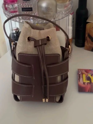 Brun och beige bucket bag i canvas - Trendig bucket bag i beige canvas med bruna läderdetaljer och vita sömmar. Väskan har justerbar axelrem och dragskostängning framtill. Perfekt för dig som gillar en cool och avslappnad stil med snygga kontraster.
