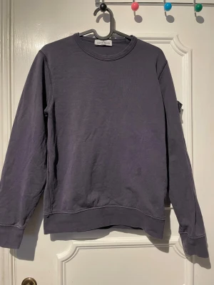 Lila/blå sweatshirt från Stone Island - Säljer en lila/blå sweatshirt från Stone Island med klassisk rund hals och ribbade muddar. Tröjan har en stilren design och den ikoniska loggan på vänster ärm. Perfekt för dig som gillar streetwear och vill ha något bekvämt och snyggt. Passar Xs/xxs