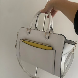 Vit handväska - Vit handväska med både handhandtag och axelrem som man kan justera id en längd man vill. Gul miniväska i ficka som detalj på framsidan (dock är dragkedjan på denna svår att stänga och öppna…). 