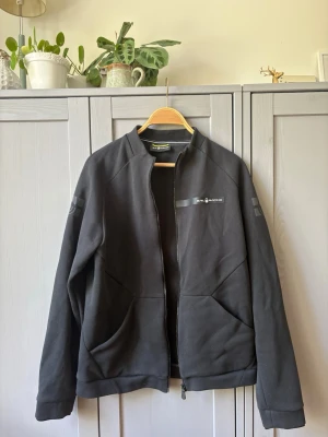 Svart hoodie från Sail Racing - Svart hoodie från Sail Racing med dragkedja framtill och två stora fickor. Diskret logga på bröstet och detaljer på ärmarna. Tillverkad i mjukt och bekvämt material, perfekt för en avslappnad stil.