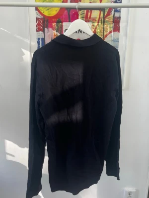 Svart tröja från Tiger of Sweden - Snygg svart långärmad tröja från Tiger of Sweden med klassisk krage och knappar framtill. Tröjan har en stilren design och är tillverkad i ett mjukt material som passar perfekt till olika outfits. Den är lite längre baktill än framtill och skulle säga att den passar storlek L också.