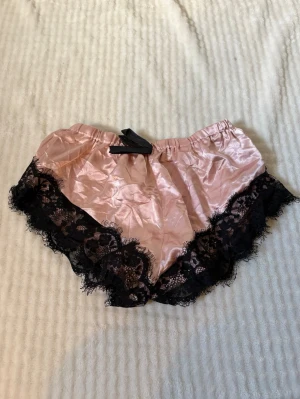 Rosa satinpyjamasshorts med spets - Snygga pyjamasshorts i glansigt rosa satin med bred svart spetskant runt benen. De har elastisk midja och en mjuk, avslappnad passform. Perfekta för mysiga hemmakvällar eller när du vill känna dig extra fin i sängen.