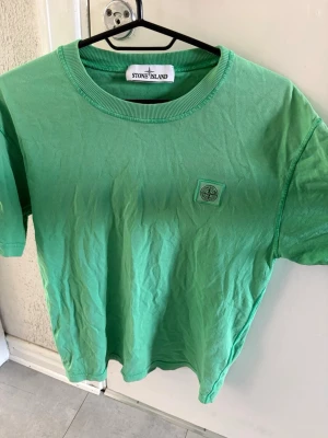 Grön t-shirt från Stone Island - Snygg grön t-shirt från Stone Island med klassisk logotyp på bröstet. T-shirten har rund halsringning och korta ärmar, perfekt för en avslappnad och trendig stil. Tillverkad i mjuk bomull som känns skön mot huden.