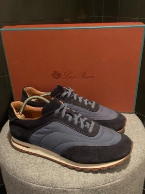 Loro Piana blå sneakers i mocka och textil - Snygga sneakers från Loro Piana i mörkblå mocka och blå textil. Skorna har klassisk låg siluett, matchande blå snörning och beige innersula i läder. Yttersulan är vit med bruna detaljer för extra stilpoäng.