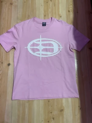 Rosa t-shirt - Snygg rosa t-shirt från Booad med en stor vit broderad logga framtill. T-shirten har klassisk passform, rund hals och är kortärmad. Perfekt för dig som gillar streetwear och vill sticka ut med en unik detalj.