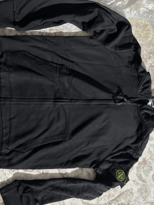 Svart hoodie från Stone Island - Svart hoodie från Stone Island med dragkedja framtill och klassisk logotyp-patch på vänster ärm. Tröjan har huva med dragsko, ribbade muddar och två stora fickor framtill. Perfekt för en stilren och avslappnad look.
