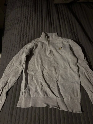 Grå stickad tröja med halvzip Lyle & Scott - Snygg grå stickad tröja från Lyle & Scott med halvzip och deras klassiska gula logga på bröstet. Tröjan har lång ärm, ribbade muddar och hög krage. Perfekt för lager-på-lager och enkel att matcha med jeans eller chinos.