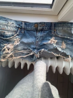 Blå slitna jeansshorts från River Island - Säljer ett par blå jeansshorts från River Island med slitna detaljer och fransig kant. Shortsen en låg midja och en cool, avslappnad look. Perfekta för sommaren. (Köpta här på plick, bara att höra av sig gällande frågor)🥰
