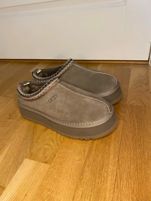 Beige UGG tofflor med platåsula - Säljer ett par beige/brun tofflor från UGG med grov platåsula och dekorativ stickning runt öppningen. Tillverkade i mocka med mjukt foder på insidan. Perfekta för att hålla fötterna varma och sköna hemma eller ute. De passar även storlek 38 