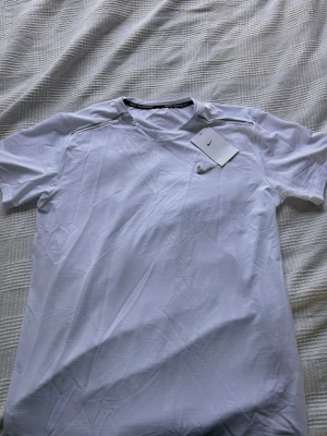 Vit Nike Dri-FIT löpartröja - Snygg vit löpartröja från Nike med Dri-FIT-teknologi som håller dig torr under träningen. Tröjan har rund hals, korta ärmar och en silverfärgad Nike-logga på bröstet. Perfekt för löpning och annan träning. Står storlek xxl men sitter som M