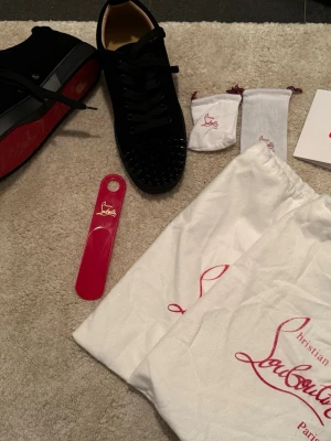 Christian Louboutin svarta sneakers - Svarta sneakers från Christian Louboutin med klassisk röd sula och svarta nitar på tån. Skorna har snörning, är låga och tillverkade i mocka och läder. Ikonisk design som sticker ut med exklusiva detaljer och tydlig logga på tillbehören.