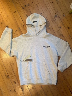 Grå Essentials Fear of God hoodie - Snygg ljusgrå hoodie från Essentials Fear of God med svart logga på bröstet och stor text på ryggen. Klassisk känguruficka framtill och stor huva. Perfekt för en avslappnad streetwear-look.