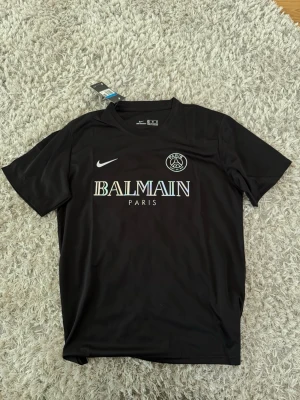 Svart PSG x Balmain tröja Nike - Snygg svart t-shirt från Nike i samarbete med PSG och Balmain. Tröjan har korta ärmar, rund hals och tryck med Balmain Paris-logga i regnbågsskimrande färg samt PSG-emblem och Nike swoosh på bröstet. Perfekt för dig som gillar streetwear och fotboll.