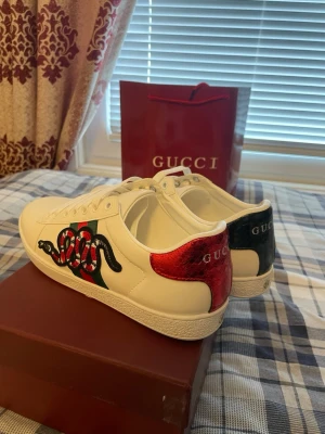 Gucci Ace sneakers med orm - Snygga vita Gucci Ace sneakers i läder med ikonisk broderad orm på sidan och klassiska gröna och röda ränder. Baksidan har metallicrött och metallicgrönt hälparti med Gucci-logga. Perfekta för dig som vill sticka ut med lyxig streetstyle.