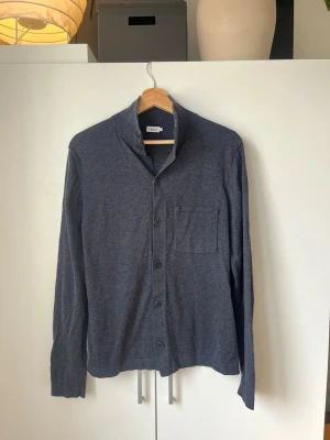 Mörkgrå cardigan från Filippa K - Stilren mörkgrå cardigan från Filippa K med knäppning framtill och en bröstficka. Tröjan har lång ärm och en mjuk, tunn kvalitet som passar perfekt för lager på lager.