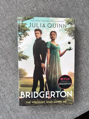 Bridgerton: The Viscount Who Loved Me - En romantisk och underhållande roman på engelska av Julia Quinn, känd från Netflix-serien Bridgerton. Boken har ett färgstarkt omslag med två personer i tidstypiska kläder och utspelar sig i historisk miljö. Perfekt för dig som gillar kärlek, drama och historiska romaner.