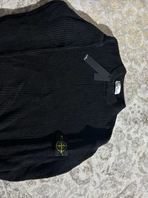 Svart stickad tröja från Stone Island - Säljer en svart stickad tröja från Stone Island med klassisk patch på ärmen. Tröjan har rund halsringning och långa ärmar med ribbade muddar. Perfekt för dig som gillar stilrena och tidlösa plagg.