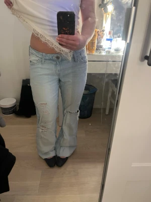 Ljusblå slitna bootcut jeans - Säljer ett par ljusblå bootcut jeans med slitningar och hål på båda benen. Jeansen har klassisk femficksdesign och låg midja. Perfekta för en avslappnad och trendig look. Upplimmade med textillim för o passa mig som är 160, innerbenslängd: ca 72cm