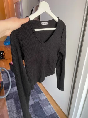 Svart ribbad långärmad topp - Svart ribbad topp från FBSister med v-ringning och långa ärmar. Modellen är croppad och har en slim passform som sitter snyggt på kroppen. Perfekt att matcha med jeans eller kjol för en trendig look.