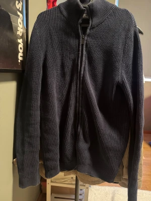 Mörkblå stickad ziptröja Dressmann - Mörkblå stickad tröja från Dressmann med dragkedja framtill och hög krage. Tröjan är ribbstickad och har långa ärmar, perfekt för lager-på-lager. Enkel och stilren design som passar till många outfits.
