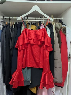 Röd volangtopp från NA-KD - Snygg röd topp från NA-KD i glansigt material med tunna axelband och volangdetaljer både framtill och på ärmarna. Perfekt för dig som vill sticka ut med en färgstark och trendig look.