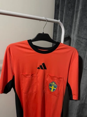 Röd domartröja från Adidas - Röd domartröja från Adidas med svarta detaljer och två bröstfickor. Tröjan har rund halsringning och svart logotyp på bröstet samt svensk fotbollsemblem. Perfekt för dig som dömer matcher eller vill ha en unik sporttröja.