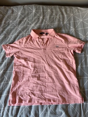 Rosa pikétröja från Gant - Klassisk rosa pikétröja från Gant med liten logga broderad på bröstet. Tröjan har krage, knappar framtill och korta ärmar. Perfekt för en avslappnad men ändå stilren look.