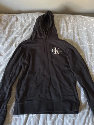 Svart hoodie från Calvin Klein - Svart hoodie från Calvin Klein med klassisk logga på bröstet. Hoodien har långa ärmar, ribbade muddar och en skön huva. Perfekt för en chill och stilren look. Materialet känns mjukt och bekvämt mot huden.