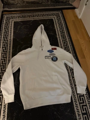 Vit hoodie från Mercier med patches - Säljer en vit hoodie från Mercier i storlek M. Hoodien har flera coola patches på bröstet och en klassisk känguruficka framtill. Den har huva med dragsko och är tillverkad i mjukt material som känns skönt mot huden. Perfekt för en avslappnad streetstyle-look.