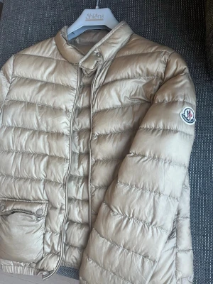 Moncler dunjacka  - En moncler dunjacka. Den är tunn så passar perfekt nu till sommaren. Färgen är guld/beige. Strl - 164 men passar mig som har S i jackor. Inga skador alls så väldigt bra kvalitet och skick.
