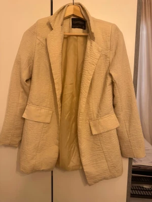 Beige kavaj från Contessa, strl 36 - Stilren beige kavaj från Contessa i storlek 36. Kavajen har en strukturerad yta, klassisk krage och två fickor framtill. Tillverkad i Turkiet och helfodrad för extra komfort. Perfekt för dig som vill ha en snygg och enkel look.