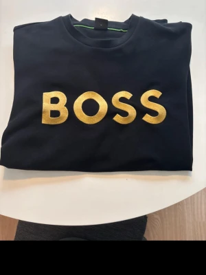 Svart hoodie från Hugo Boss - Svart hoodie från Hugo Boss med stor guldfärgad BOSS-logga på bröstet. Den är som ny och säljer för den ej passar kostar endast 650 men vid snabbt köp kan jag tänka mig att gå ner lite i pris