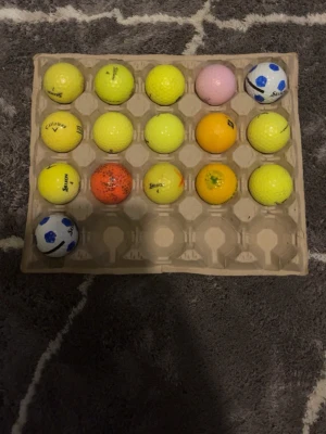 Färgglada golfbollar i blandade märken - Säljer ett set med 15 golfbollar i olika färger som gult, orange, rosa och vitt med blå detaljer. Bollarna är av olika märken som Callaway och Srixon och ligger i en äggkartong. Perfekt för dig som vill ha extra bollar till golfträningen.
