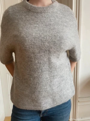 Ljusgrå stickad t-shirt - Mysig ljusgrå stickad t-shirt med korta ärmar och rund hals. Tröjan har en avslappnad passform och är tillverkad i ett mjukt, fluffigt material som känns skönt mot huden. Perfekt att styla med jeans eller kjol för en enkel och trendig look.