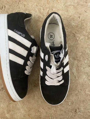 Adidas Campus svarta sneakers - Svarta Adidas Campus sneakers med klassiska vita ränder på sidorna och vit sula med gummidetalj. Ovandel i mocka och vita skosnören. Ikonisk retrostil med låg profil och tydlig Adidas-logga på plösen.