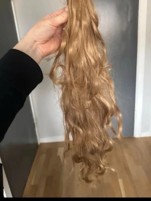 Lång lockig hårförlängning blond - Säljer en lång, lockig hårförlängning i en naturlig blond nyans. Perfekt för dig som vill ha extra volym och längd till håret. Hårdelen är syntetisk och har en mjuk, glansig finish med fina lockar.