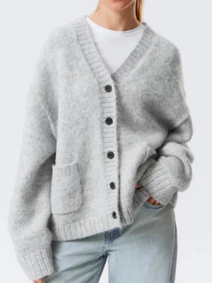 Grå stickad kofta från Weekday - Mysig gråmelerad kofta från H&M med oversized passform, djup V-ringning och stora svarta knappar framtill. Koftan har ribbade muddar, en framficka och är stickad i ett mjukt material som känns fluffigt och skönt.