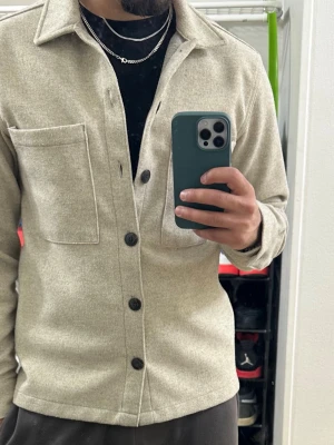 Beige overshirt från Jack & Jones - Stilren beige overshirt från Jack & Jones med klassisk krage, två stora bröstfickor och svarta knappar framtill. Tillverkad i ett mjukt, ull-liknande material som ger en clean och modern look. Perfekt att bära över en t-shirt för en avslappnad stil.