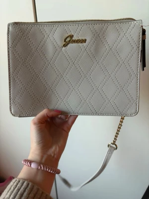 Beige quiltad handväska från Guess - Snygg beige handväska från Guess med quiltat rutmönster och guldfärgad logga framtill. Väskan har avtagbar axelrem och detaljer i både läder och kedja. Perfekt storlek för det viktigaste och stängs med dragkedja. Stilren och trendig accessoar.