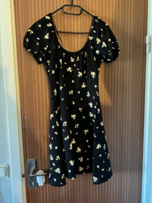 Svart blommig kortklänning med puffärm - Supersöt svart kortklänning med små blommor i gult och vitt. Klänningen har puffärmar och en rundad urringning. Perfekt för varmare dagar och ger en romantisk vibe. Materialet känns lätt och flowy.