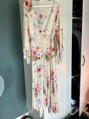 Blommig långklänning från Stajl  - Romantisk långklänning från By Malina i ljus bas med blommigt mönster i rosa, blått och grönt. Klänningen har v-ringning, vida ärmar, volangdetaljer och ett knytband i midjan. Perfekt för sommarens alla tillfällen.