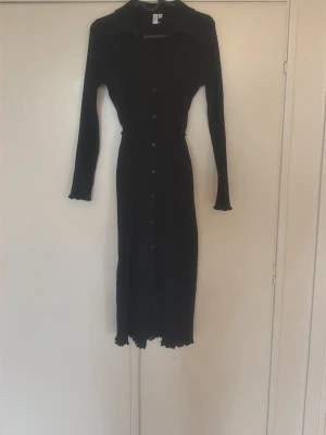 Svart långärmad klänning med knappar - Svart långklänning med långa ärmar och knappar längs hela framsidan. Klänningen har en krage, smal passform och ett bälte i midjan. Materialet är ribbat och stretchigt, vilket ger en snygg siluett. Perfekt för dig som gillar stilrena och klassiska plagg.