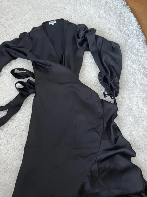 Svart omlottklänning i satin - Supersnygg svart omlottklänning från  bubbleroomi glansigt satinmaterial. Klänningen har långa puffärmar med knyt vid handlederna och markerad midja med knytband i sidan. Perfekt för dig som gillar en elegant och trendig look.