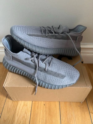 Grå Yeezy Boost 350 V2 sneakers - Snygga Yeezy Boost 350 V2 i helgrå färg med stickad ovandel och genomskinlig rand på sidan. Skorna har en chunky, räfflad sula och klassiska snörningar. Perfekta för dig som gillar streetwear och vill ha något unikt på fötterna.