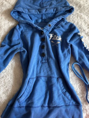 Blå hoodie från Abercrombie & Fitch  - Snygg blå hoodie från Abercrombie & Firvv med broderad logga på bröstet och längs ärmen. Hoodien har knappar framtill, magficka och huva. Snörena till huvan har dragits ur, men det är bara att stoppa tillbaka dem (om man vill så gör jag gärna det, men säg då till). Står storlek M, men passar nog mer Xs. Inga synbara defekter förutom lite lösa trådar som syns i bilden. 
