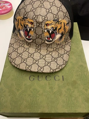 Gucci keps med tigertryck - Snygg keps från Gucci med klassiskt GG-mönster i beige och brunt samt två stora tigerhuvuden framtill. Kepsen har böjd skärm och mesh på sidorna för extra ventilation. Perfekt för dig som vill sticka ut med lyxig streetstyle.